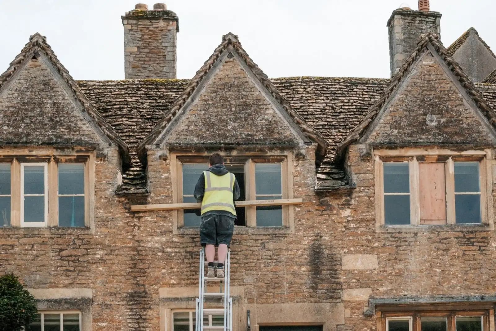 Builder in hi-vis on a ladder repairing a window on a stone-built English house