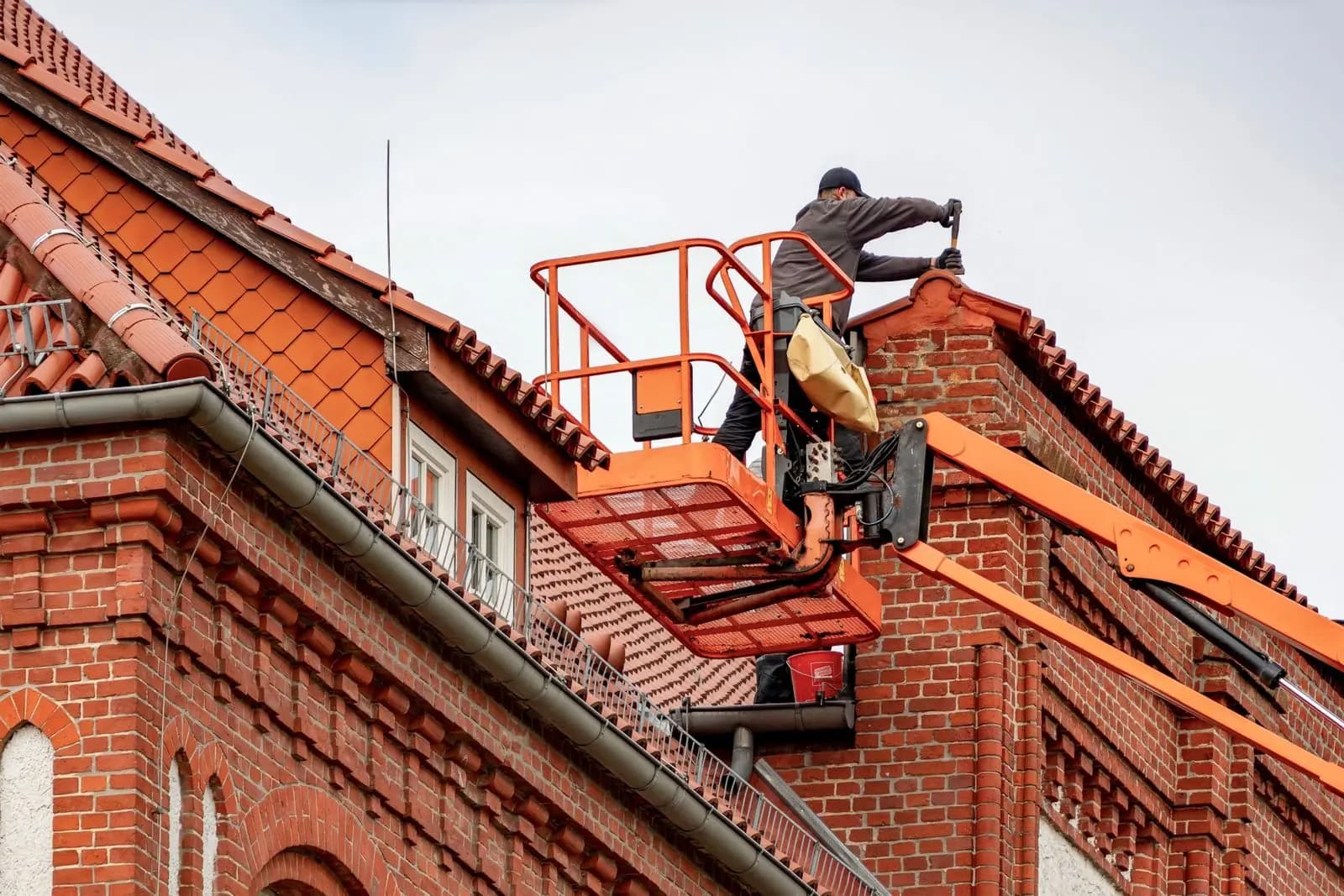 Roofer on a cherry picker repairing the clay-tiled roof of a red-brick building