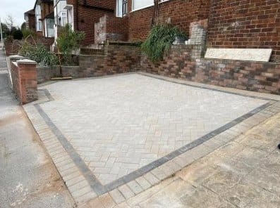 Light grey herringbone block-paved driveway with a darker border, in front of a brick terrace
