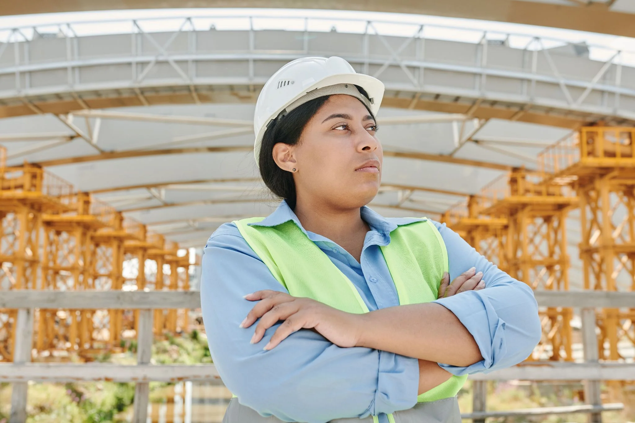 A female construction manager wearing a white hard hat and a high-visibility safety vest stands confidently with her arms crossed at a large infrastructure construction site.