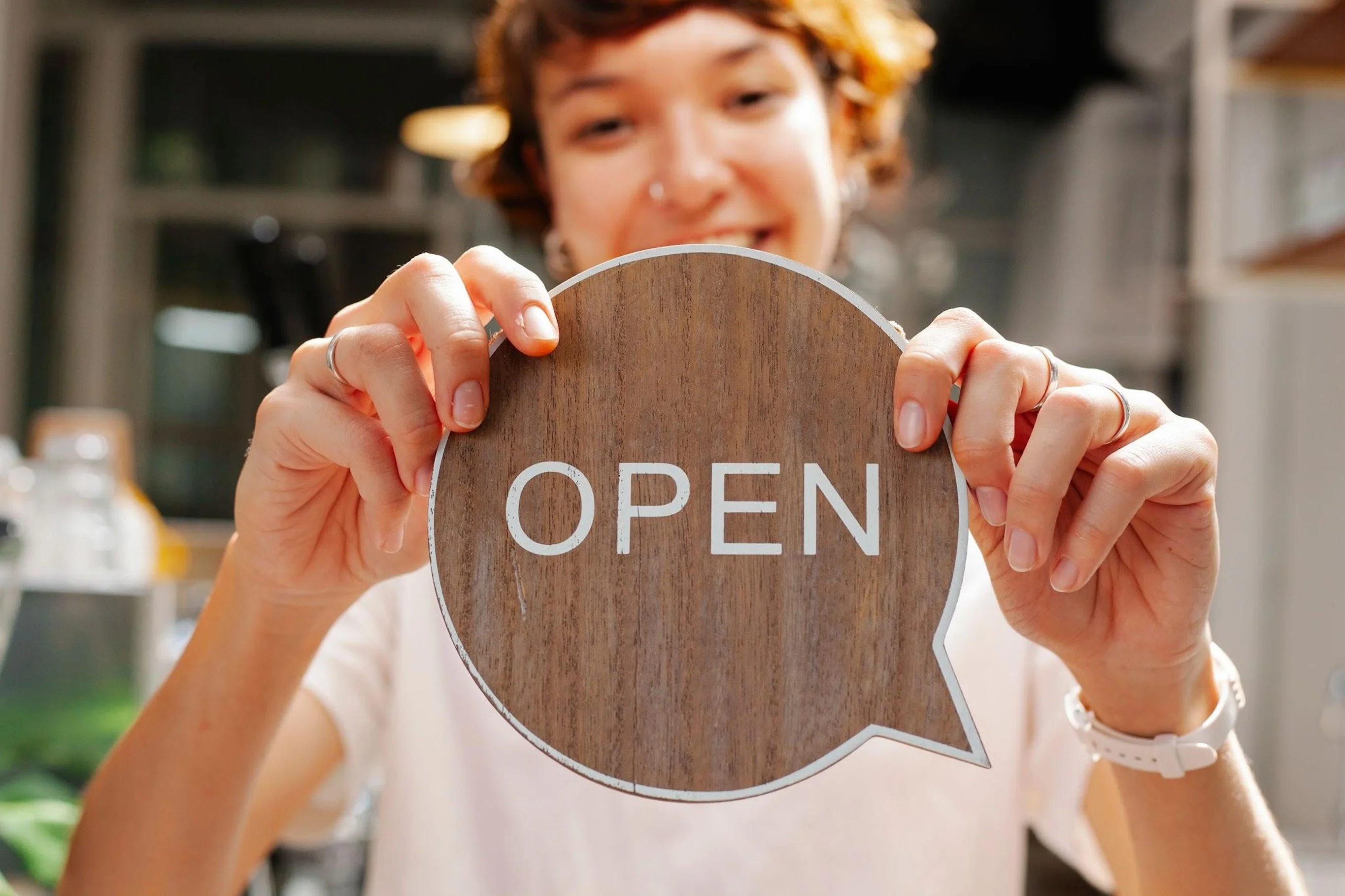 A smiling small business owner holding a wooden speech bubble sign with the word "OPEN" printed on it.