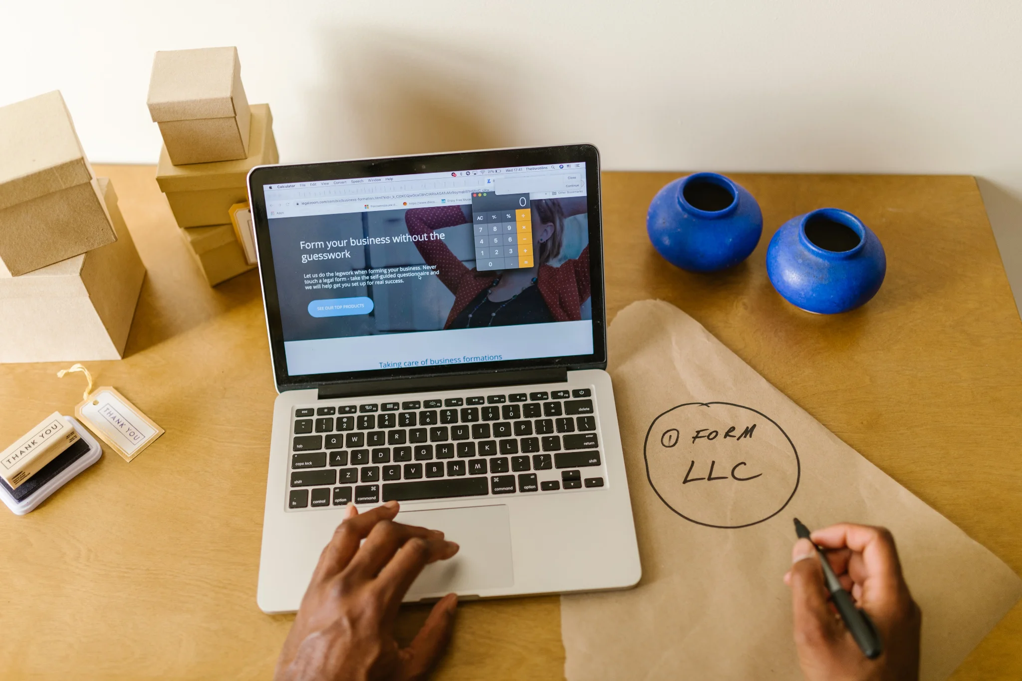 Small business owner planning company setup and writing LLC formation notes next to a laptop displaying digital tools and packaging supplies.